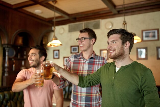 people, men, leisure, friendship and celebration concept - happy male friends clinking beer glasses and watching sport game or football match at bar or pub