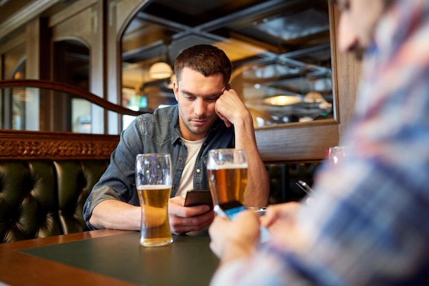 people, men, leisure, friendship and communication concept - male friends with smartphones drinking draft beer at bar or pub