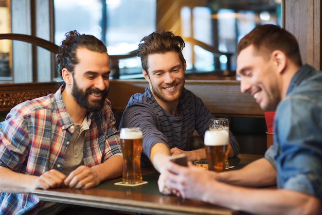 people, men, leisure, friendship and technology concept - happy male friends with smartphone drinking beer at bar or pub