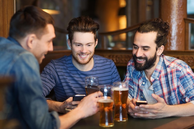 people, men, leisure, friendship and technology concept - happy male friends with smartphones drinking beer at bar or pub
