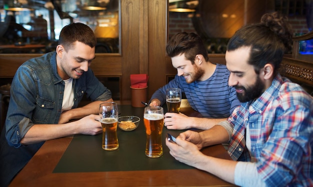 people, men, leisure, friendship and technology concept - male friends with smartphones drinking beer at bar or pub