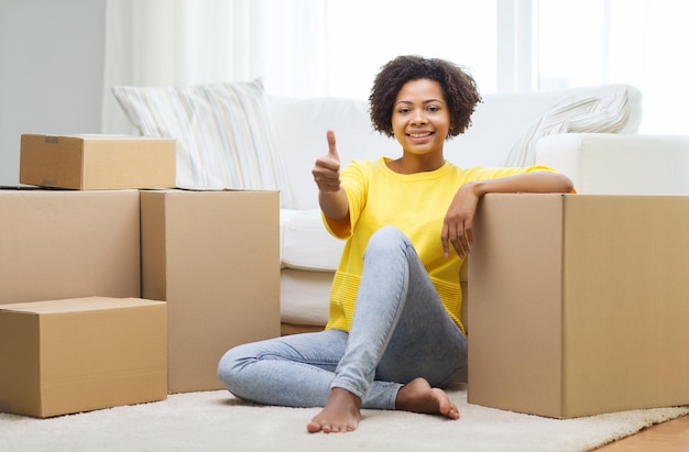 people, moving new place, gesture and repair concept - happy african american young woman with many cardboard boxes sitting on floor and showing thumbs up at home