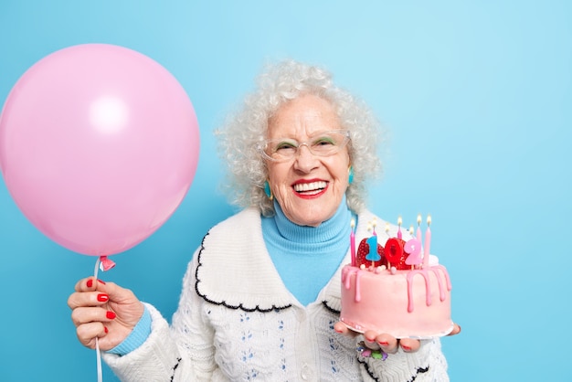 People old age partying and holidays concept. Positive beautiful grandmother dressed in neat clothes celebrates her 102nd birthday holds inflated balloon and tasty cake