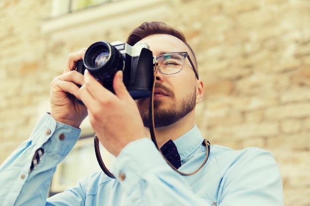 people, photography, technology, leisure and lifestyle - happy young hipster man with retro vintage film camera taking picture on city street