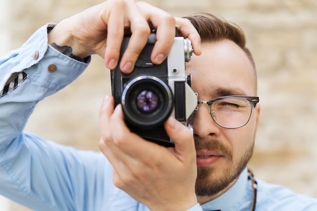 people, photography, technology, leisure and lifestyle - happy young hipster man with retro vintage film camera taking picture on city street