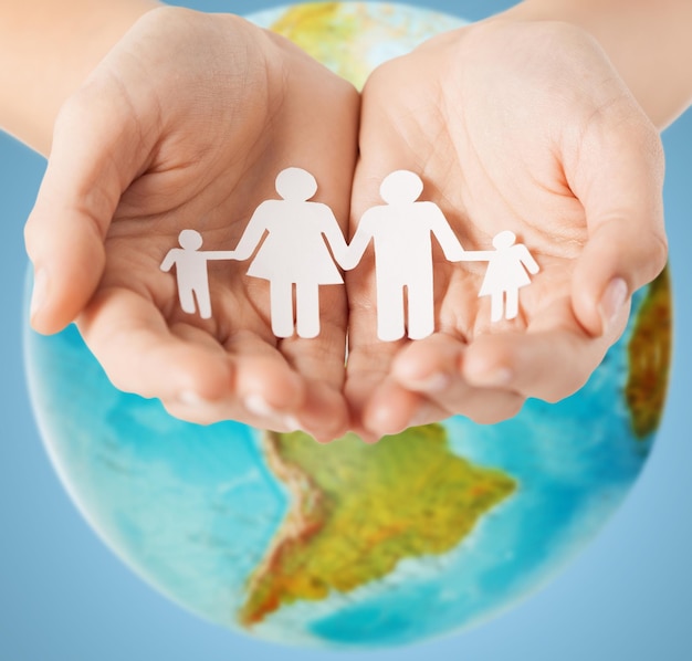 people, population, charity and life concept - close up of human hands holding paper family over earth globe and blue background