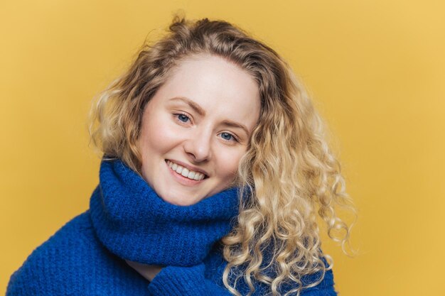 People positive emotions and beauty concept Beautiful lovely young smiling female model in blue warm sweater being satisfied with something has pleasant smile on face isolated on yellow wall