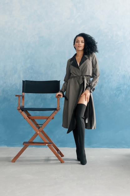 People, race, ethnicity and fashion concept - beauty black model posing in high boots. Young pretty beautiful mulatto woman sitting in director's chair at film. woman in gray raincoat, afro hairstyle