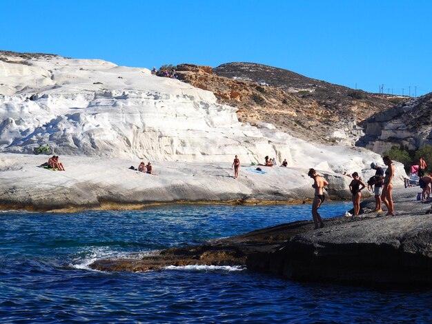 People on rocks by mountains against clear sky