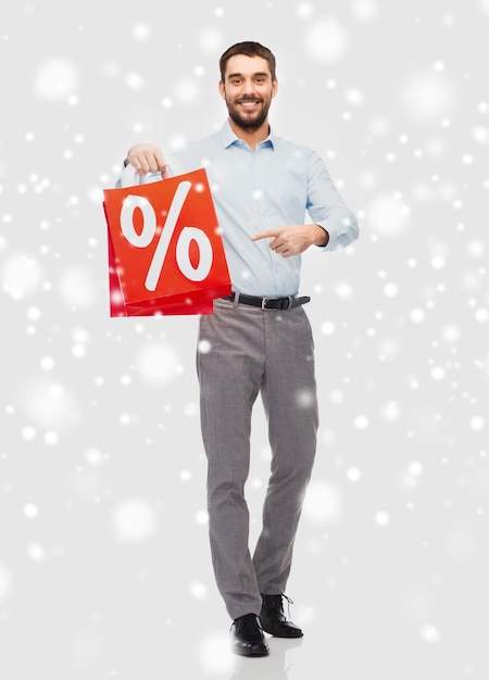 people, sale, christmas, winter and holidays concept - smiling man holding red shopping bag with percentage sign over snow background