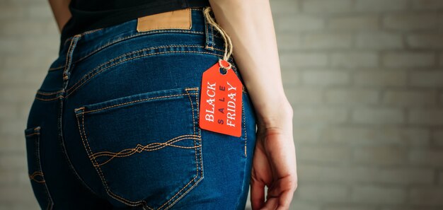 Persone, vendita, concetto di consumismo. chiuda sulla foto ritagliata delle natiche della donna sessuale che indossa pantaloni in denim casual blu su sfondo grigio mattone nel centro commerciale. etichetta rossa venerdì nero sui jeans.