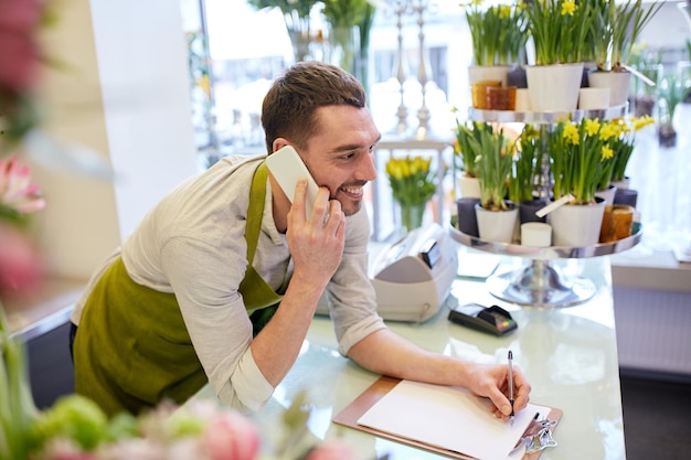 people, sale, retail, business and floristry concept - happy smiling florist man calling on smartphone and making notes to clipboard at flower shop counter