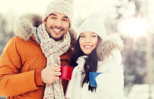 people, season, love, drinks and leisure concept - happy couple holding hot tea cups over winter landscape