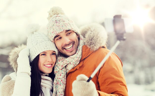 people, season, love, technology and leisure concept - happy couple taking picture with smartphone selfie stick on over winter background