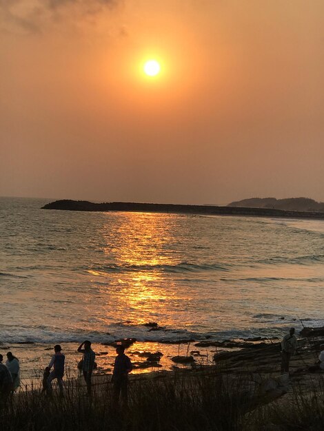 People on shore at beach against orange sky