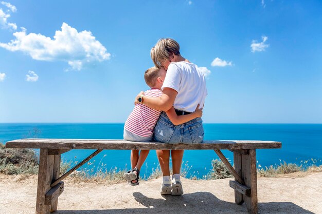 People sit on a wooden bench with a magnificent view of the sea on a sunny day Mom with a little son in tshirts and shorts hug and kiss Active lifestyle and travel Back view