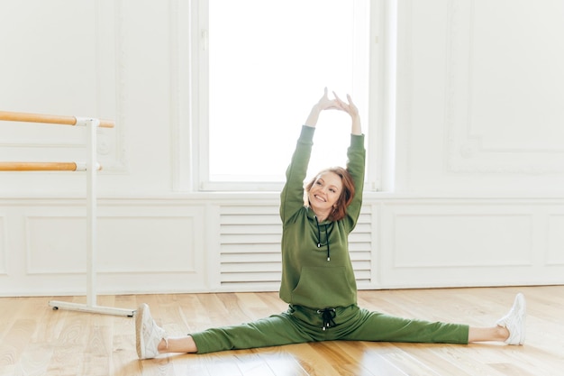 People strength fitness gymnastics concept Glad sportswoman with cheerful face raises hands spreads legs and does splits stretches in studio Young dancer has work out dressed in tracksuit