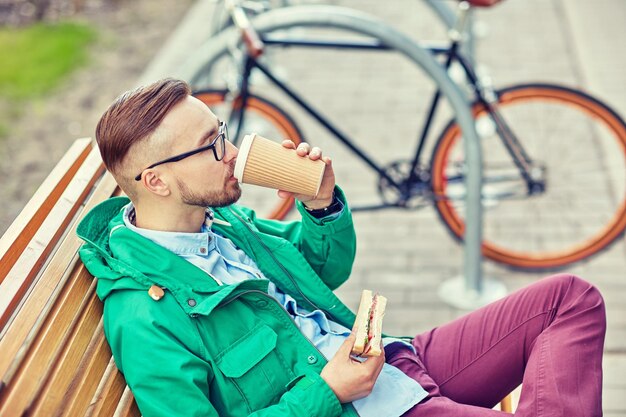 people, style, leisure and lifestyle - happy young hipster man drinking coffee cup and eating sandwich on city street