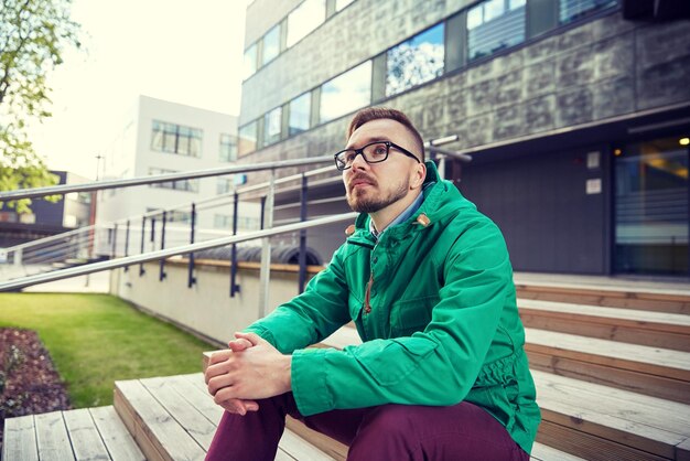 people, style, leisure and lifestyle - happy young hipster man in eyeglasses and jacket sitting on stairs in city