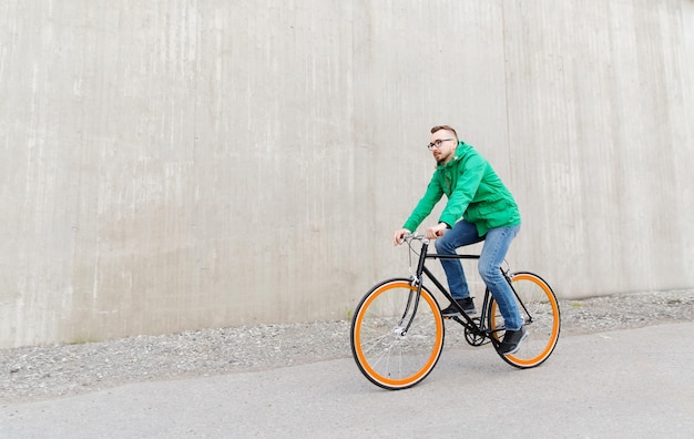 people, style, leisure and lifestyle - happy young hipster man riding fixed gear bike on city street