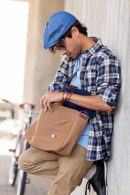 people, style, leisure and lifestyle - hipster man lookir for something in his shoulder bag and fixed gear bike on city street