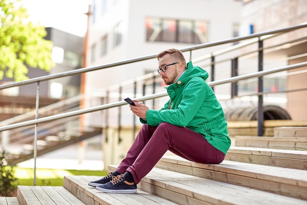 Photo people, style, technology and lifestyle - young hipster man with smartphone sitting on stairs in city