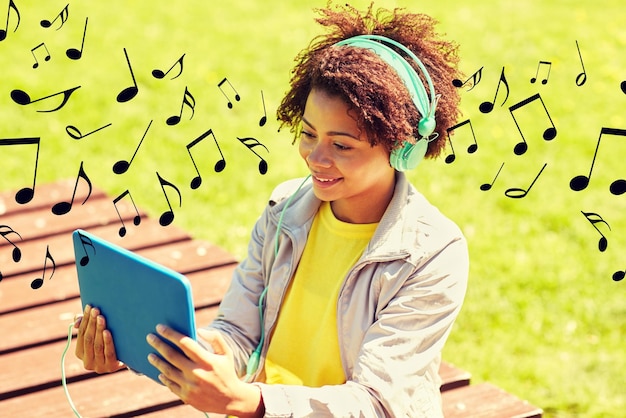 people, summer, technology and leisure concept - happy african american young woman in headphones with tablet pc computer listening to music or watching video outdoors over notes background