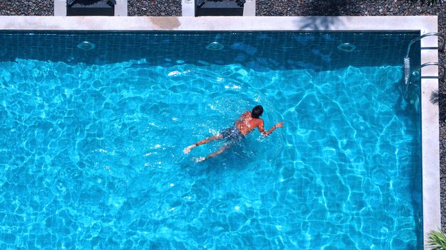 Люди плавают в бассейне под углом обзора сверху, вода синего цвета и солнечный свет отражаются на поверхности.