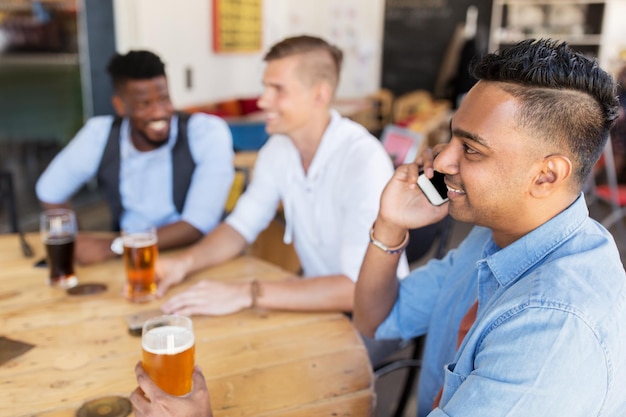 Photo people, technology and communication concept - happy man calling on smartphone and drinking beer with friends at bar or pub
