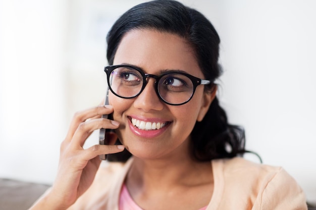 人、テクノロジー、コミュニケーション、レジャーのコンセプト – 自宅でスマートフォンで通話する眼鏡をかけた幸せな笑顔の若いインド人女性
