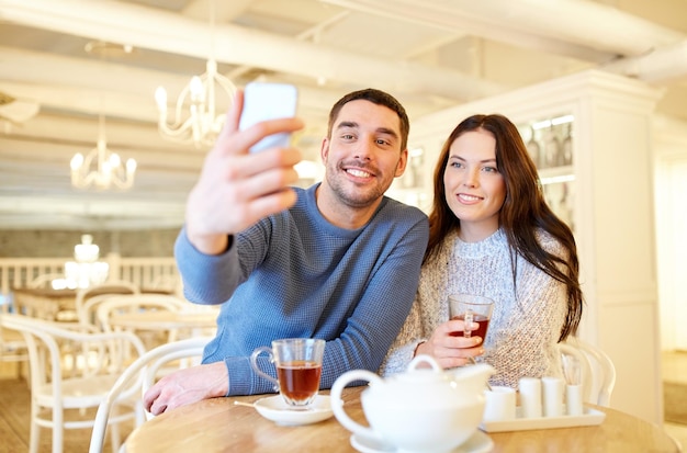 people, technology and dating concept - happy couple taking smartphone selfie and drinking tea at cafe or restaurant