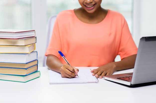 people, technology, learning and education concept - close up of happy african american young woman sitting at table with laptop computer and books and writing to notebook at home