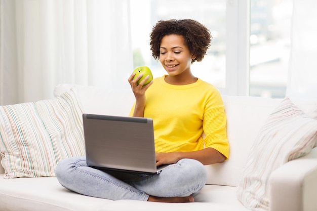 people, technology and leisure concept - happy african american young woman sitting on sofa with laptop computer and drinking tea from cup at home