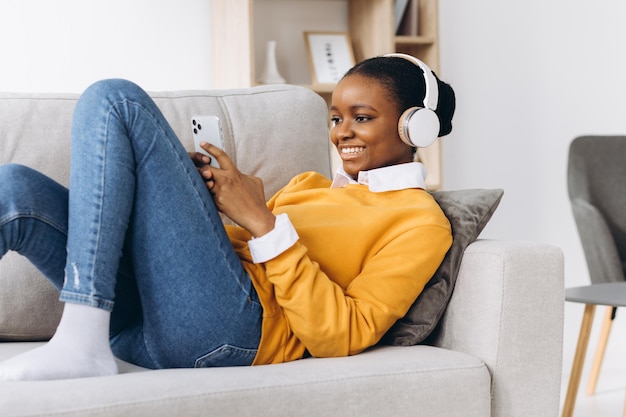 People, technology and leisure concept - happy african american young woman sitting on sofa with smartphone and headphones listening to music at home