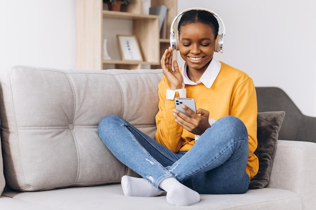 사람, 기술, 레저 개념 - 스마트폰과 헤드폰을 들고 소파에 앉아 집에서 음악을 듣는 행복한 아프리카계 미국인 젊은 여성