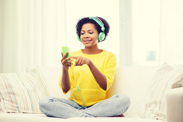 people, technology and leisure concept - happy african american young woman sitting on sofa with smartphone and headphones listening to music at home