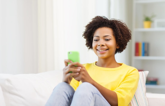 people, technology and leisure concept - happy african american young woman sitting on sofa with smartphone at home