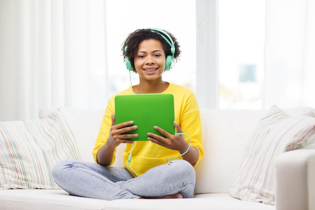 人、テクノロジー、レジャーのコンセプト – 幸せなアフリカ系アメリカ人の若い女性がソファに座り、タブレットPCコンピュータとヘッドホンで家で音楽を聴く
