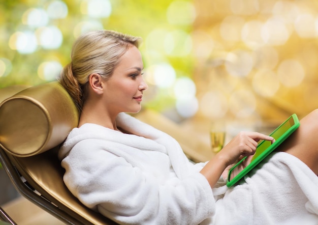 people, technology and relaxation concept - beautiful young woman in white bath robe with tablet pc computer social networking at spa over holidays lights background