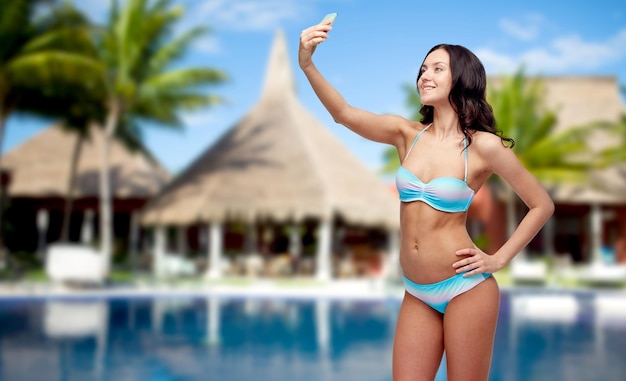 people, technology, summer vacation and travel concept - happy young woman in bikini swimsuit taking selfie with smatphone over swimming pool, bungalow and palm trees at hotel resort background