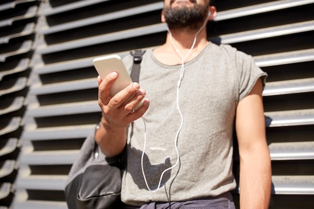人、テクノロジー、旅行、観光のコンセプト – 路上で音楽を聴くイヤホン、スマートフォン、バッグを持つ男性の接写