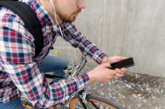 people, travel, technology, leisure and lifestyle - close up of young hipster man in earphones with smartphone and fixed gear bike listening to music on city street