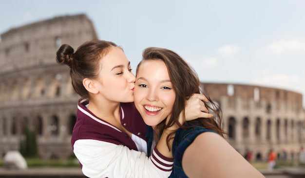 people, travel, tourism and friendship concept - happy smiling pretty teenage girls taking selfie and kissing over coliseum in rome background