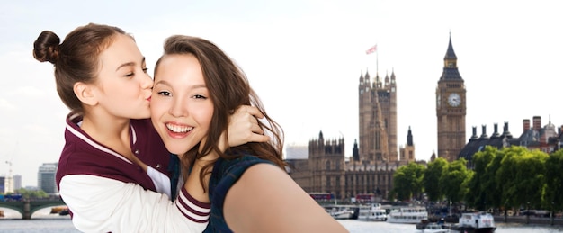 people, travel, tourism and friendship concept - happy smiling pretty teenage girls taking selfie and kissing over london background
