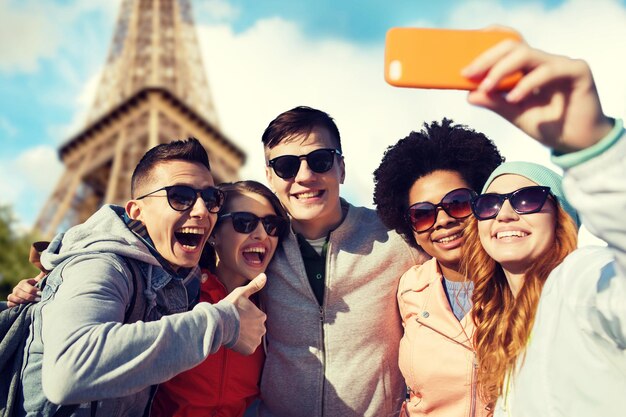 people, travel, tourism, friendship and technology concept - group of happy teenage friends taking selfie with smartphone and showing thumbs up over paris eiffel tower background