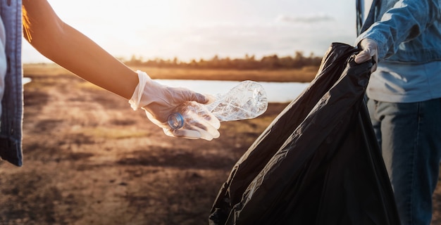 People volunteer keeping garbage plastic bottle into black bag at park near river in sunset