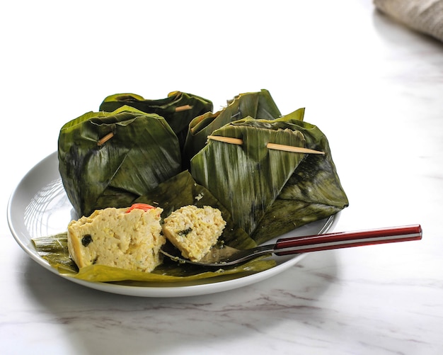 Pepes tahu is indonesian spiced tofu wrapped with banana leaf\
and steamed, typically indonesian food from west java (sundanese).\
steamed tofu with asian basil, white background