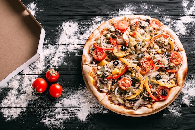 Foto pizza ai peperoni accanto a una scatola per il confezionamento di farina versata su un tavolo di legno nero