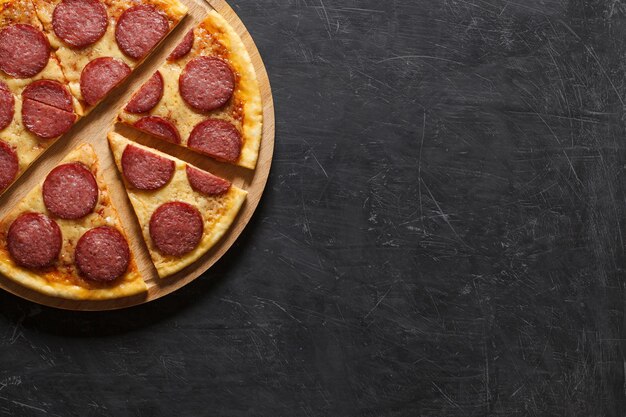 Pepperoni pizza cut into pieces on cutting wooden board on dark background with space to copy text