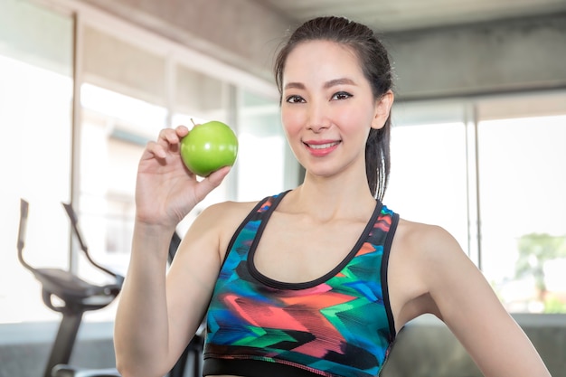 Совершенная азиатская женщина в sportswear держа зеленое яблоко для ест перед разминкой на спортзале фитнеса.